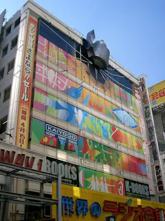 A colorful building in Japan features large, bright murals with abstract designs and Japanese text. The facade displays "KAIYODO Hobby Lobby Tokyo" and "K-BOOKS" signs, with a capsule toy above the entrance.