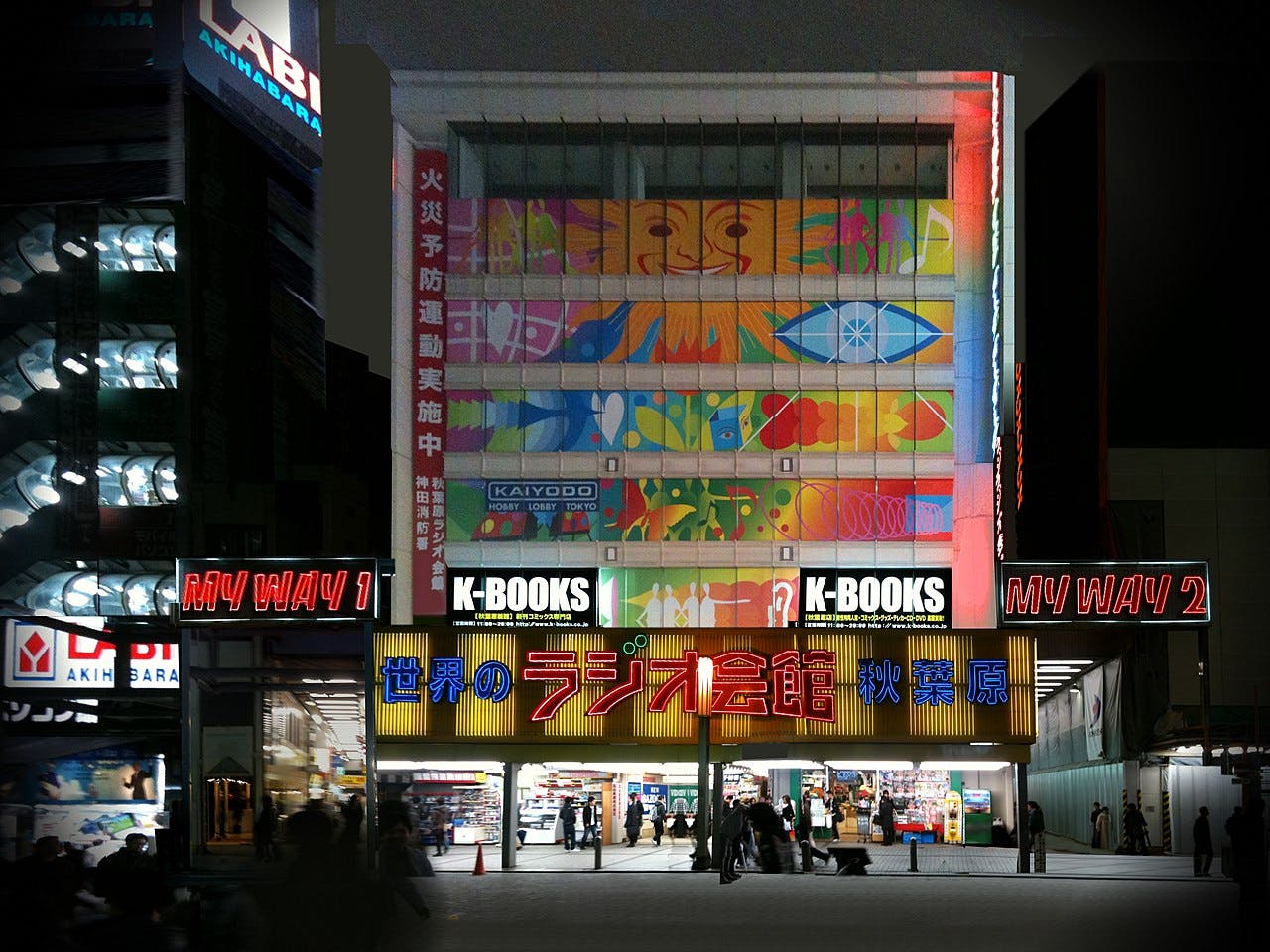 A brightly lit multi-story building at night in Akihabara, Tokyo, displays colorful signs in Japanese and English, including "K-BOOKS" and "RADIO KAIKAN," with people walking in front.