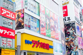 Colorful city buildings in Japan covered with bright anime posters, billboards, and advertisements in Japanese text, showcasing animated characters and pop culture imagery.