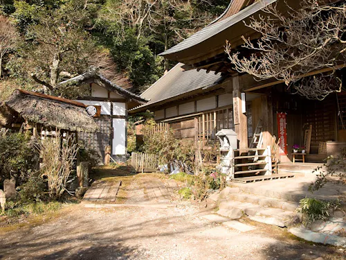 Amida-ji A traditional Japanese shrine complex with wooden buildings, one with a thatched roof and another with tiled roofing, surrounded by trees and plants on a sunny day. Steps and stone lanterns are visible in the foreground.