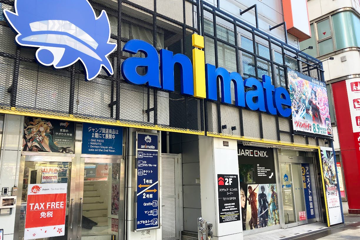 Storefront of Animate, a popular Japanese anime and manga shop. The exterior features large blue and yellow signage, anime posters, and promotional banners. The entrance displays “TAX FREE” and other advertisements.