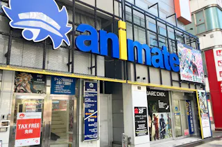 Storefront of Animate, a popular Japanese anime and manga shop. The exterior features large blue and yellow signage, anime posters, and promotional banners. The entrance displays “TAX FREE” and other advertisements.