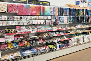 Store shelves display colorful stationery items, including notebooks, folders, pens, stickers, and gift bags. Brightly designed products are neatly organized in rows, with labels and signage in Japanese visible above the items.