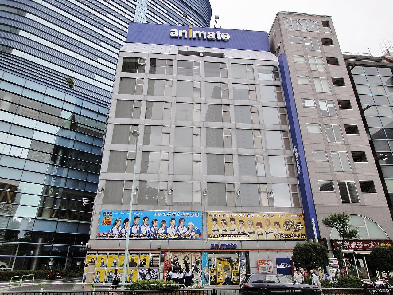 A multi-story building with a large "animate" sign at the top. The ground floor and facade display colorful posters of anime characters. Adjacent modern buildings are visible, and people are walking on the sidewalk in front.