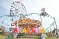 A colorful "ARAKAWA" sign stands in front of a merry-go-round, with a Ferris wheel and a small monorail ride overhead, set in an amusement park on a bright, sunny day.