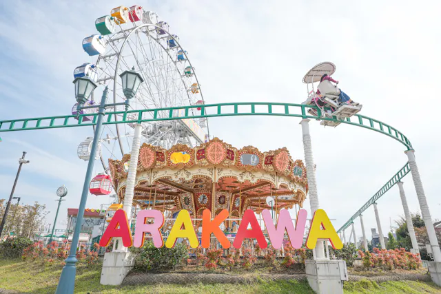 A colorful "ARAKAWA" sign stands in front of a merry-go-round, with a Ferris wheel and a small monorail ride overhead, set in an amusement park on a bright, sunny day.