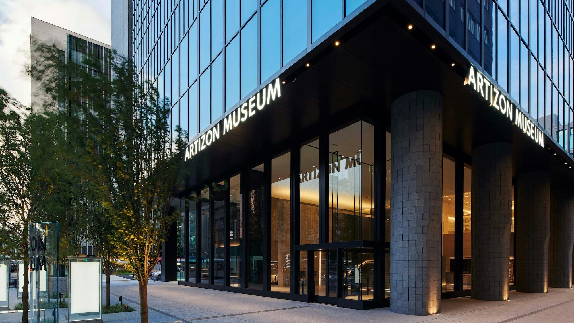 The exterior of the Artizon Museum with glass walls, tall black columns, and a modern entrance. Trees and a city street are visible beside the building. The museum name is displayed in large white letters.