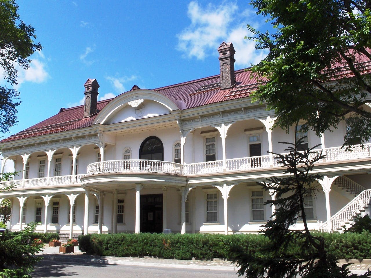 A large white colonial-style building with a red roof, two chimneys, an arched entrance, and a wraparound balcony, surrounded by trees and greenery under a bright blue sky.