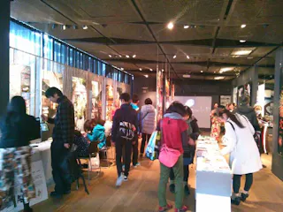 A group of people explore an indoor exhibition space with bright lighting, tables displaying items, and colorful artwork on the walls. Some visitors are seated, while others stand and look at displays.