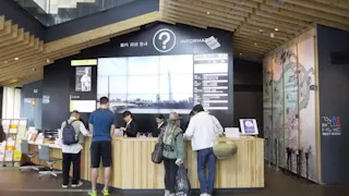 Five people stand at an information counter inside a modern building, with a large display screen showing a cityscape and a question mark symbol above. Wooden ceiling beams and a decorative wall are visible.