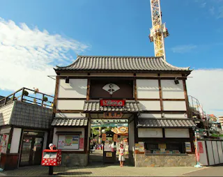 Traditional Japanese-style entrance gate with tiled roof at an amusement park, signs in Japanese, and a tall yellow tower ride visible in the background under a blue sky with clouds. Visitors are walking nearby.