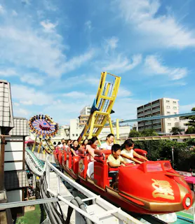 A red roller coaster filled with people speeds along a track at an outdoor amusement park, with colorful rides and buildings visible in the background under a bright blue sky.