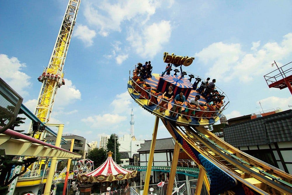 People ride a spinning disk roller coaster at an amusement park on a sunny day, with a tall yellow drop tower, carousel, and other attractions visible in the background.