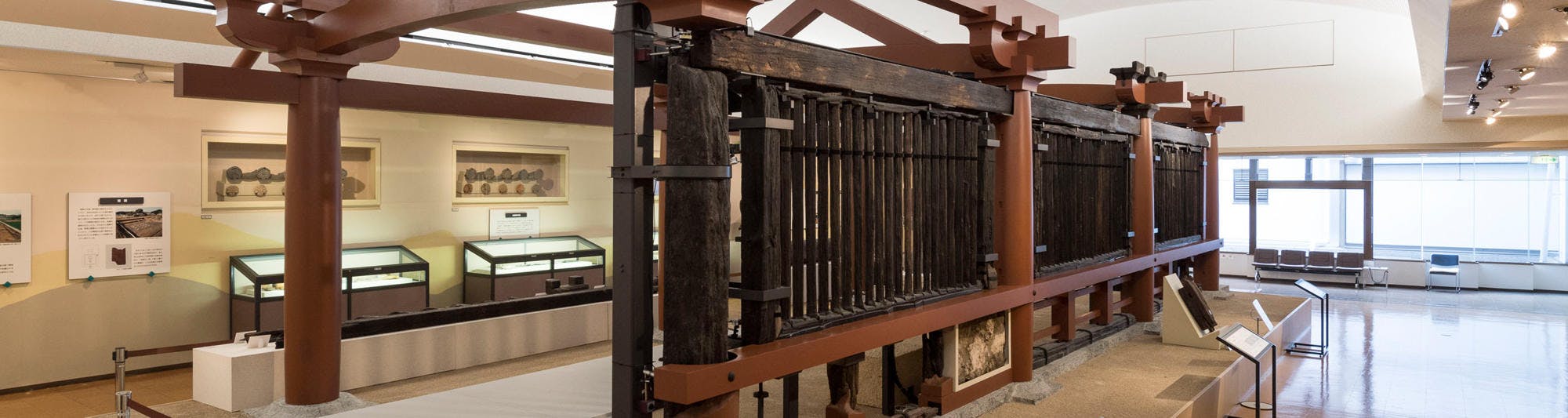 A large, wooden and metal historical gate is displayed in a museum with informational signs, glass display cases, and paintings on the walls in a well-lit, spacious room.