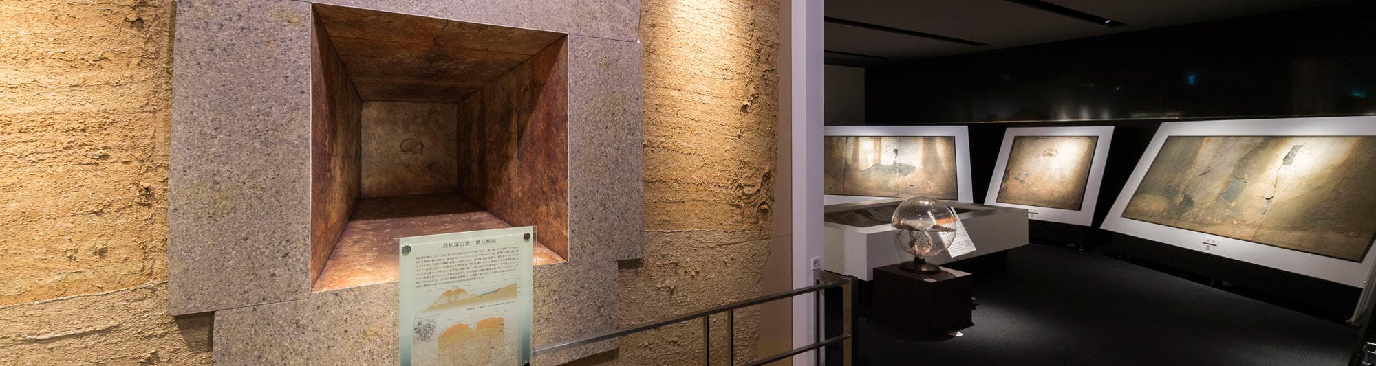 Exhibit of ancient tomb murals in a museum, featuring a recessed stone chamber on the left and brightly lit framed murals displayed on the right in a dimly lit room.