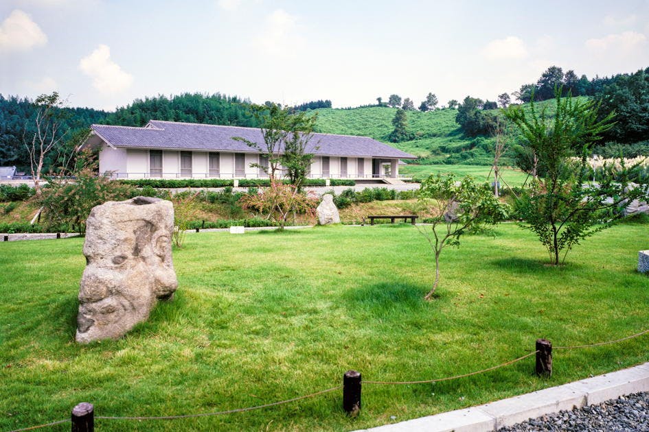 A tranquil Japanese-style garden with neatly trimmed grass, large rocks, small trees, and a traditional building in the background, set against a lush green hillside under a partly cloudy sky.