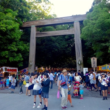 Atsuta Festival A large crowd gathers under a traditional wooden torii gate at a festival, surrounded by green trees and food stalls. Many people, including children, are enjoying snacks and wearing summer clothing.