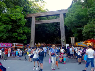 A large crowd gathers under a traditional wooden torii gate at a festival, surrounded by green trees and food stalls. Many people, including children, are enjoying snacks and wearing summer clothing.