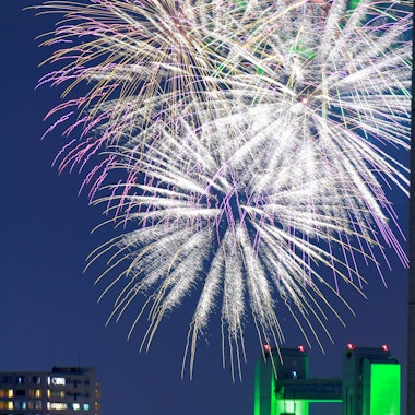Atsuta Festival Large, colorful fireworks burst in the night sky above a cityscape with tall buildings, some lit in green and white lights. The sky is dark blue, and the fireworks dominate the upper part of the image.