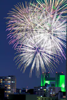 Large, colorful fireworks burst in the night sky above a cityscape with tall buildings, some lit in green and white lights. The sky is dark blue, and the fireworks dominate the upper part of the image.