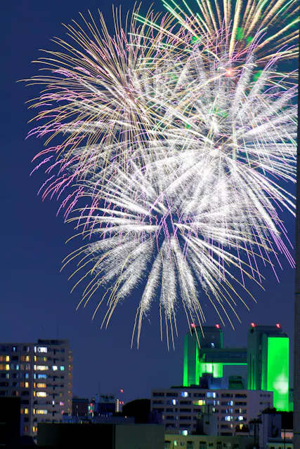 Large, colorful fireworks burst in the night sky above a cityscape with tall buildings, some lit in green and white lights. The sky is dark blue, and the fireworks dominate the upper part of the image.