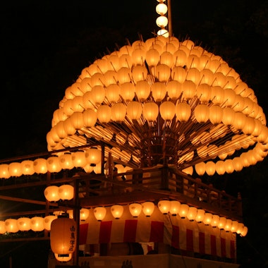 Atsuta Festival A large wooden festival float covered in rows of glowing orange paper lanterns is illuminated at night, surrounded by darkness. The float has a striped red and white lower section and a hanging lantern on one corner.