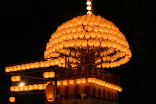 A large wooden festival float covered in rows of glowing orange paper lanterns is illuminated at night, surrounded by darkness. The float has a striped red and white lower section and a hanging lantern on one corner.
