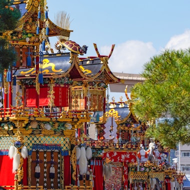 Autumn Takayama Festival (Hachiman Matsuri) Colorful, ornate Japanese festival floats decorated with gold, red, and intricate patterns are surrounded by green trees, set against a bright blue sky and modern buildings in the background.