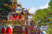 Colorful, ornate Japanese festival floats decorated with gold, red, and intricate patterns are surrounded by green trees, set against a bright blue sky and modern buildings in the background.