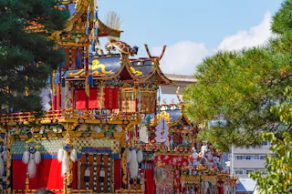Colorful, ornate Japanese festival floats decorated with gold, red, and intricate patterns are surrounded by green trees, set against a bright blue sky and modern buildings in the background.