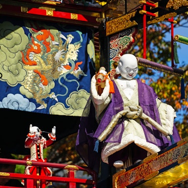Autumn Takayama Festival (Hachiman Matsuri) A colorful festival float features a large, white-faced doll in traditional Japanese attire and a smaller doll nearby, set against an ornate backdrop with a tiger and clouds design. Autumn leaves are visible in the background.