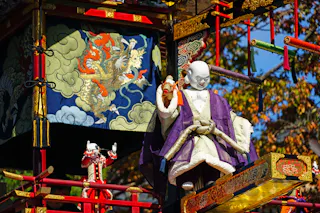 A colorful festival float features a large, white-faced doll in traditional Japanese attire and a smaller doll nearby, set against an ornate backdrop with a tiger and clouds design. Autumn leaves are visible in the background.