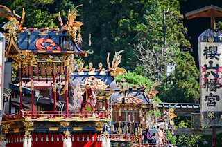 Ornate, colorful Japanese festival floats with gold and red decorations are paraded outdoors, surrounded by lush green trees and traditional wooden buildings. A large vertical sign with Japanese characters is visible on the right.