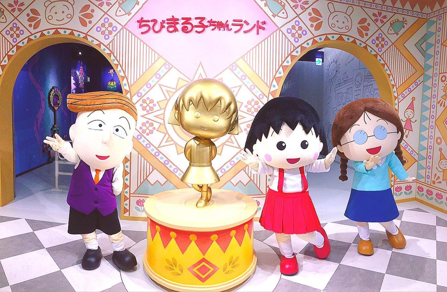 Three costumed characters pose around a gold statue of a girl in a red-and-yellow-themed room, with Japanese text and colorful decorations on the wall behind them.
