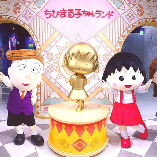 Chibi Maruko Chan Land Three costumed characters pose around a gold statue of a girl in a red-and-yellow-themed room, with Japanese text and colorful decorations on the wall behind them.