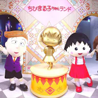 Chibi Maruko Chan Land Three costumed characters pose around a gold statue of a girl in a red-and-yellow-themed room, with Japanese text and colorful decorations on the wall behind them.
