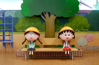Two cartoon girl statues wearing yellow hats and school uniforms sit on a bench in front of a mural depicting a tree, a fence, and animal drawings in a colorful indoor setting.