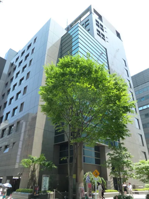 A modern mid-rise building with reflective glass windows and stone facade, partially obscured by a tall, leafy green tree in the foreground on a bright sunny day.