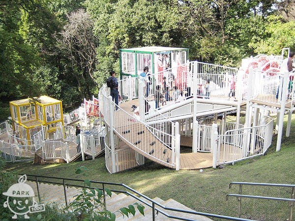 Children and adults play on a large, multi-level outdoor climbing structure with white railings and platforms, set on a grassy slope surrounded by trees. Stairs lead down to the play area.