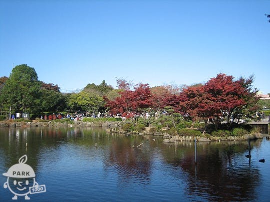 A tranquil pond reflects clear blue sky and trees with green and red foliage in a Japanese garden. People walk in the background, enjoying the scenery. A "Park" logo is in the bottom left corner.