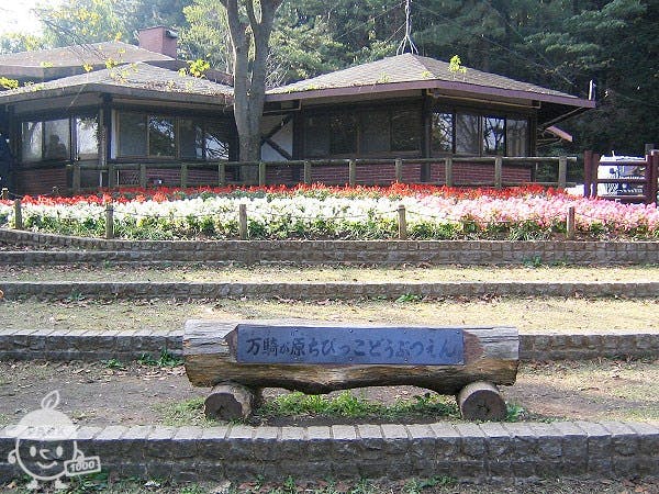 A rustic building stands behind a flower bed filled with red and white blooms. In front, a wooden sign with Japanese writing rests on a low stone step, surrounded by trees and greenery.