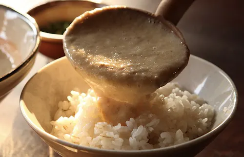 A wooden ladle pours thick, creamy grated yam sauce (tororo) over a bowl of steamed white rice, with another dish visible in the background.