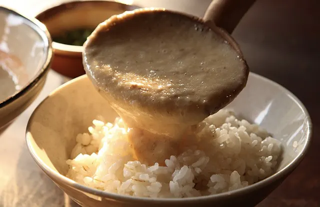 A wooden ladle pours thick, creamy grated yam sauce (tororo) over a bowl of steamed white rice, with another dish visible in the background.