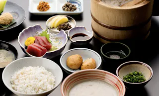 A traditional Japanese meal set with steamed rice, sashimi, pickles, dipping sauces, two fried balls, chopped green onions, miso soup, and assorted side dishes arranged neatly on a table.