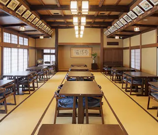 A traditional Japanese dining room with low wooden tables and chairs on tatami mats, shoji screen windows, wooden ceiling beams, and framed artwork lining the upper walls. Warm lighting creates a serene atmosphere.