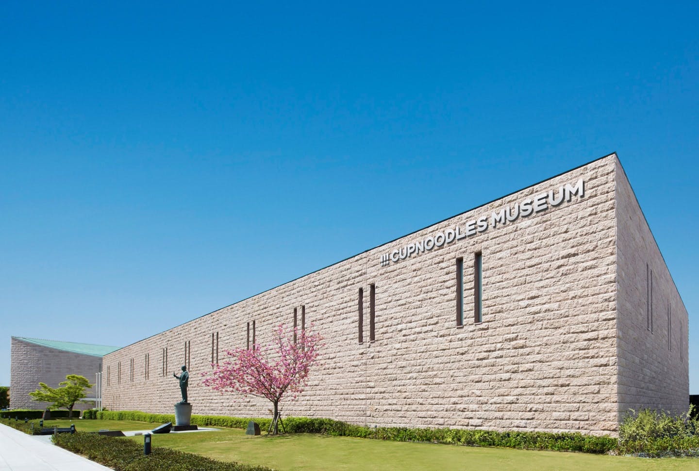 A modern, light stone building with "CUPNOODLES MUSEUM" written on the side, surrounded by green grass, a sculpted tree, and a clear blue sky.