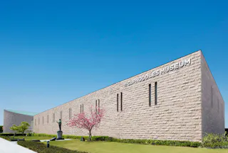A modern, light stone building with "CUPNOODLES MUSEUM" written on the side, surrounded by green grass, a sculpted tree, and a clear blue sky.