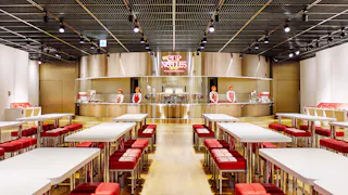 A modern, brightly-lit dining area with white tables and red cushioned benches, facing a stainless steel counter labeled "Cup Noodles," where staff in uniforms stand behind the counter.