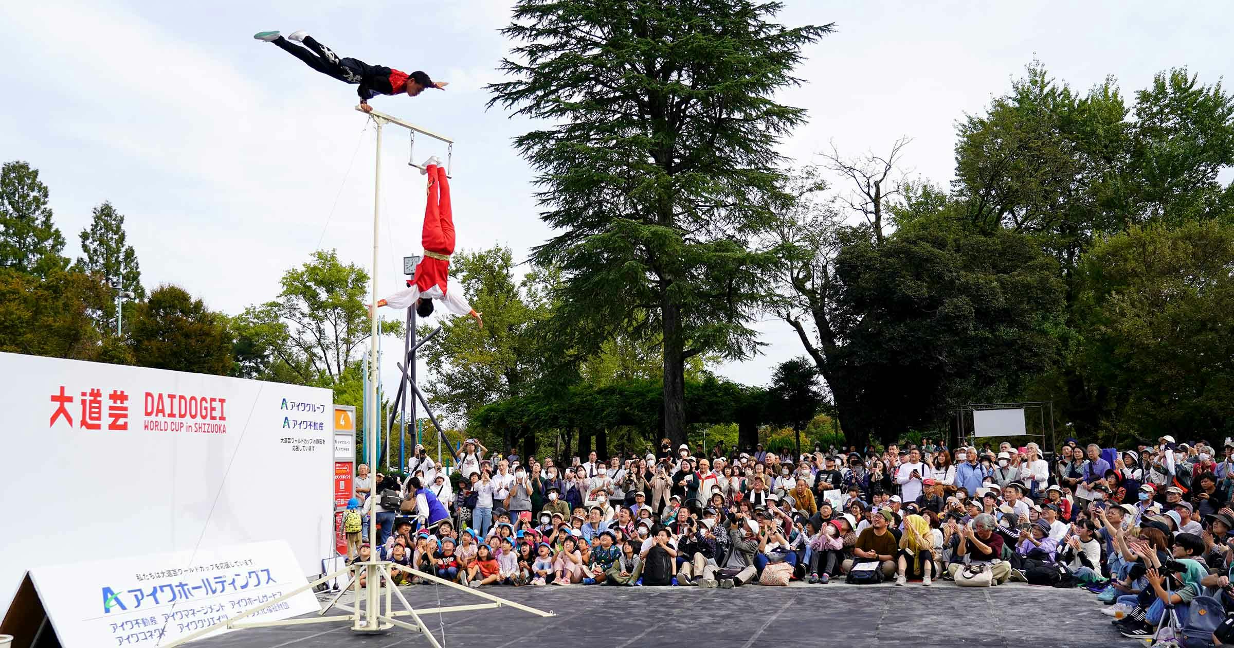 A large crowd watches as two acrobats perform; one hangs upside down on a tall pole while the other is captured mid-air in a dramatic leap above them, set outdoors with trees in the background.