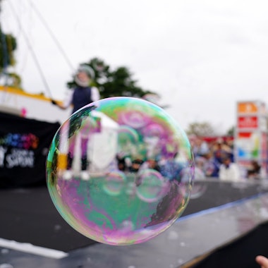 Daidogei World Cup A large, iridescent soap bubble floats in sharp focus at an outdoor event, with people, colorful tents, and a stage blurred in the background.